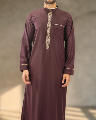 A man wearing a deep plum Saudi thobe with band collar, geometric placket embroidery, and gold piping trim.