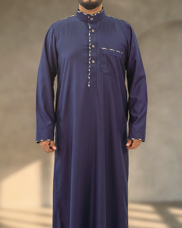 A man wearing a navy blue Saudi thobe with geometric pattern trim along the collar, placket, chest pocket, and shoulders.