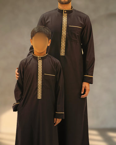 Men's black Saudi thobe featuring gold intricate embroidery on the front placket with gold piping on the collar and cuffs, shown with matching boys thobe.