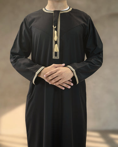 Men's black Emirati kandura with gold embroidery trim, traditional chest tassels, and long sleeves with embroidered cuffs.